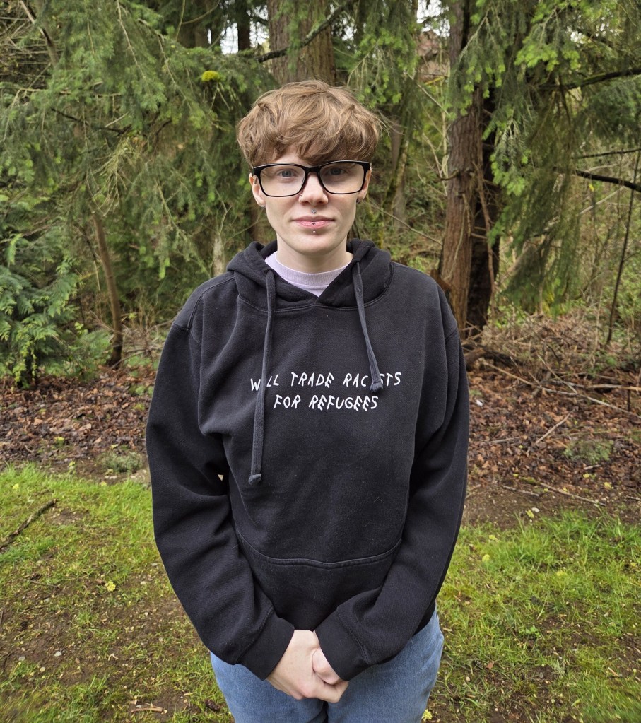 White nonbinary person with short blond hair, glasses, and a black sweater that reads in white letters, "Will trade racists for refugees". They are standing with their hands together in front of them in front of green and brown forest scenery.